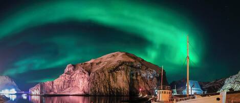 Un bote llamado "VE 21" atracado en una costa rocosa con un edificio grande e iluminado al fondo y la aurora boreal en el cielo.