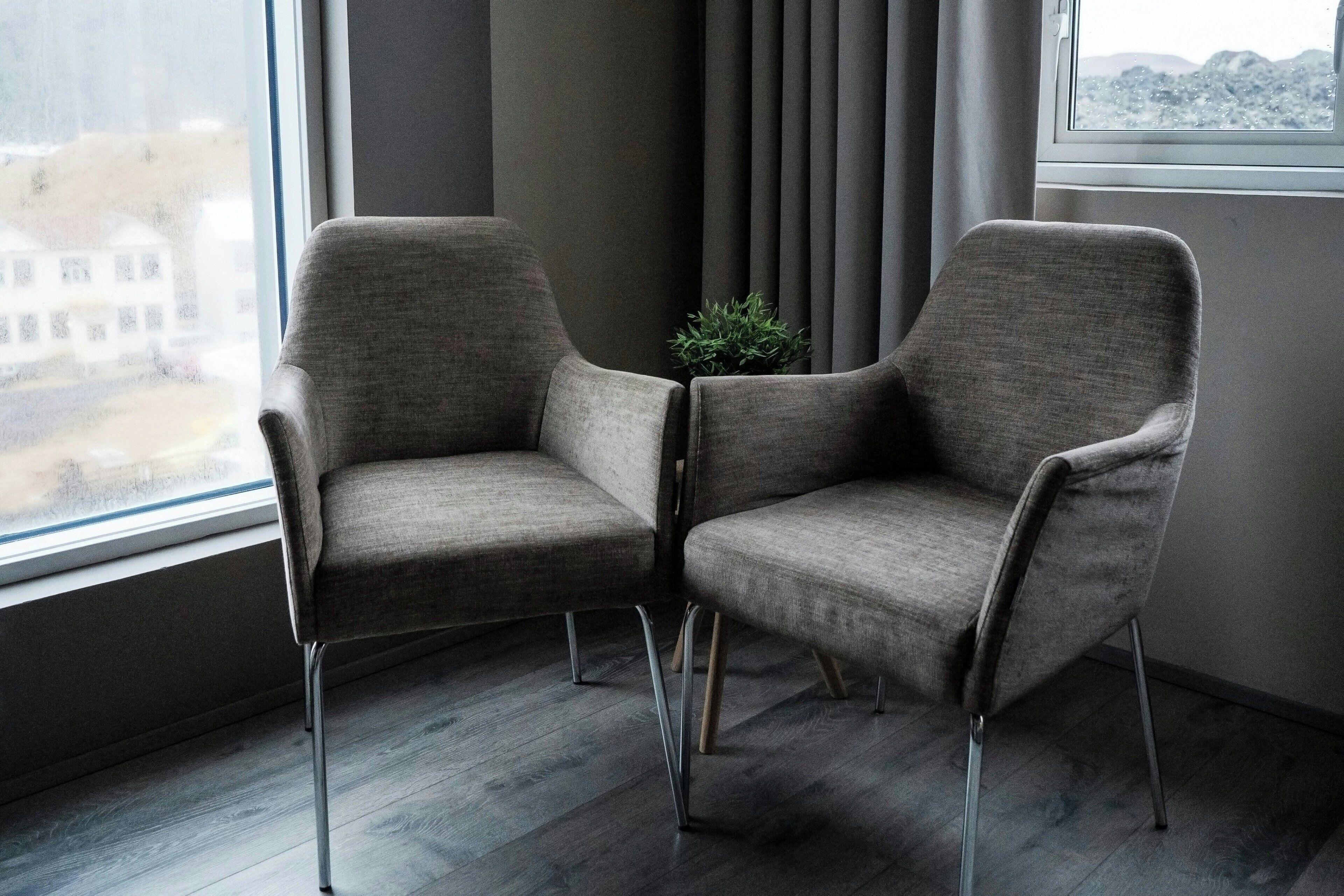 Dos sillones grises con patas de metal en una habitación con un ventanal grande, una planta y vistas a un edificio.