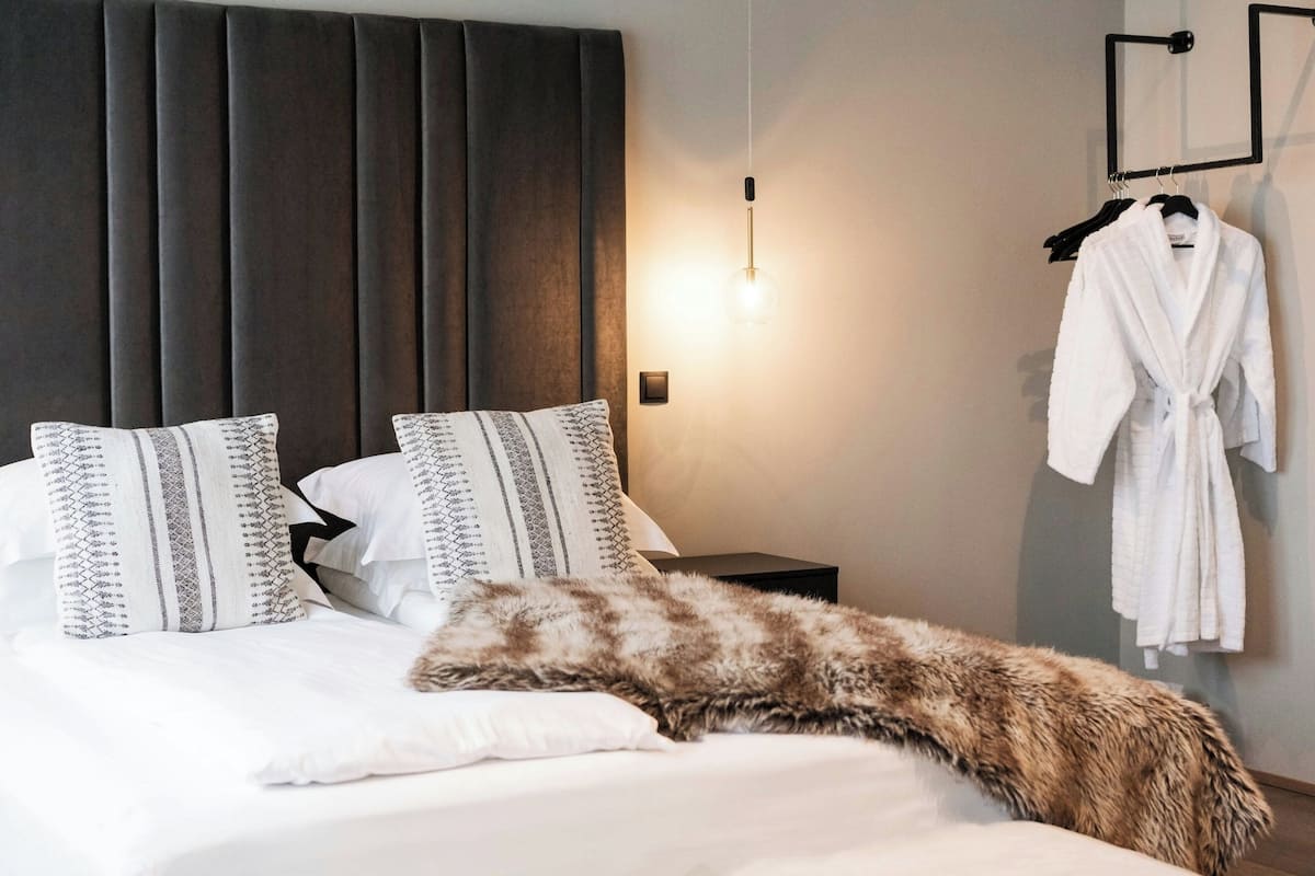 Ein ordentlich bezogenes Bett mit gemusterten Kissen, einer flauschigen Tagesdecke, einem dunklen Kopfteil, einer Nachttischlampe und einem Bademantel, der an einem Gestell hängt.