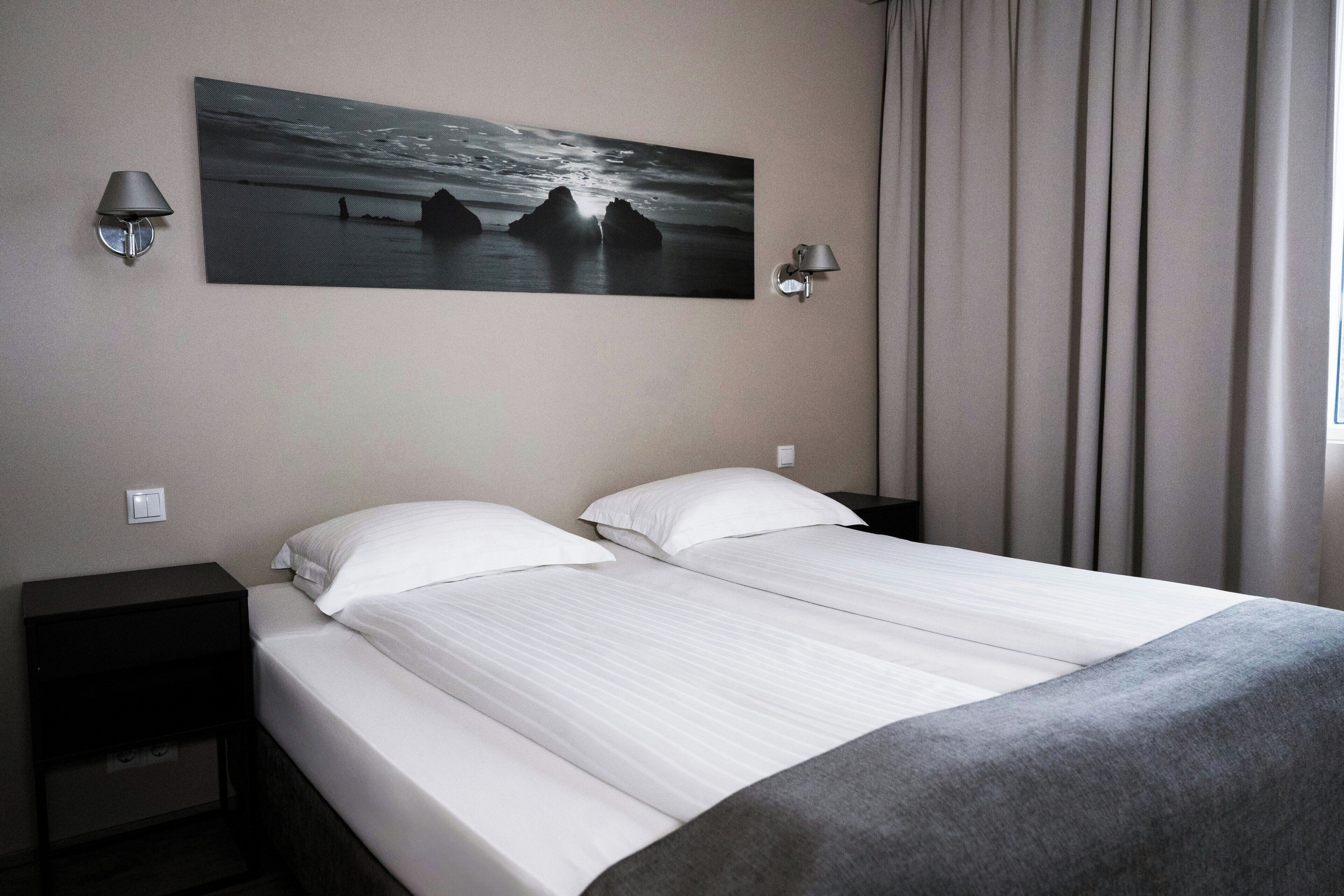 A double bed with white linens, two bedside tables with lamps, a painting of a seascape, and a window with curtains.
