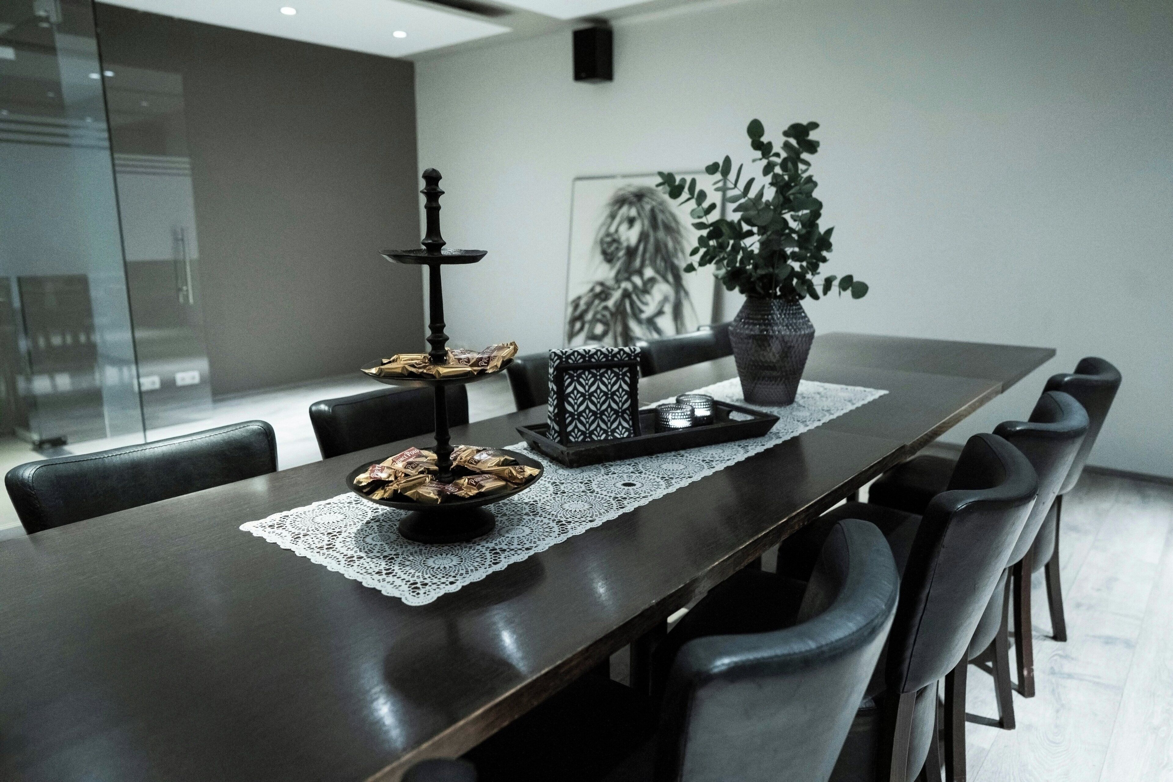 A modern dining area with a long table, black chairs, a lace table runner, a tray with food, a candle holder, and a potted plant.