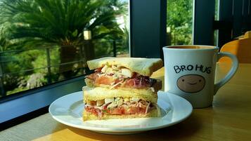 A sandwich on a plate next to a mug with a smiling face and the text "BRO HUG" on a table by a window with a view of greenery.