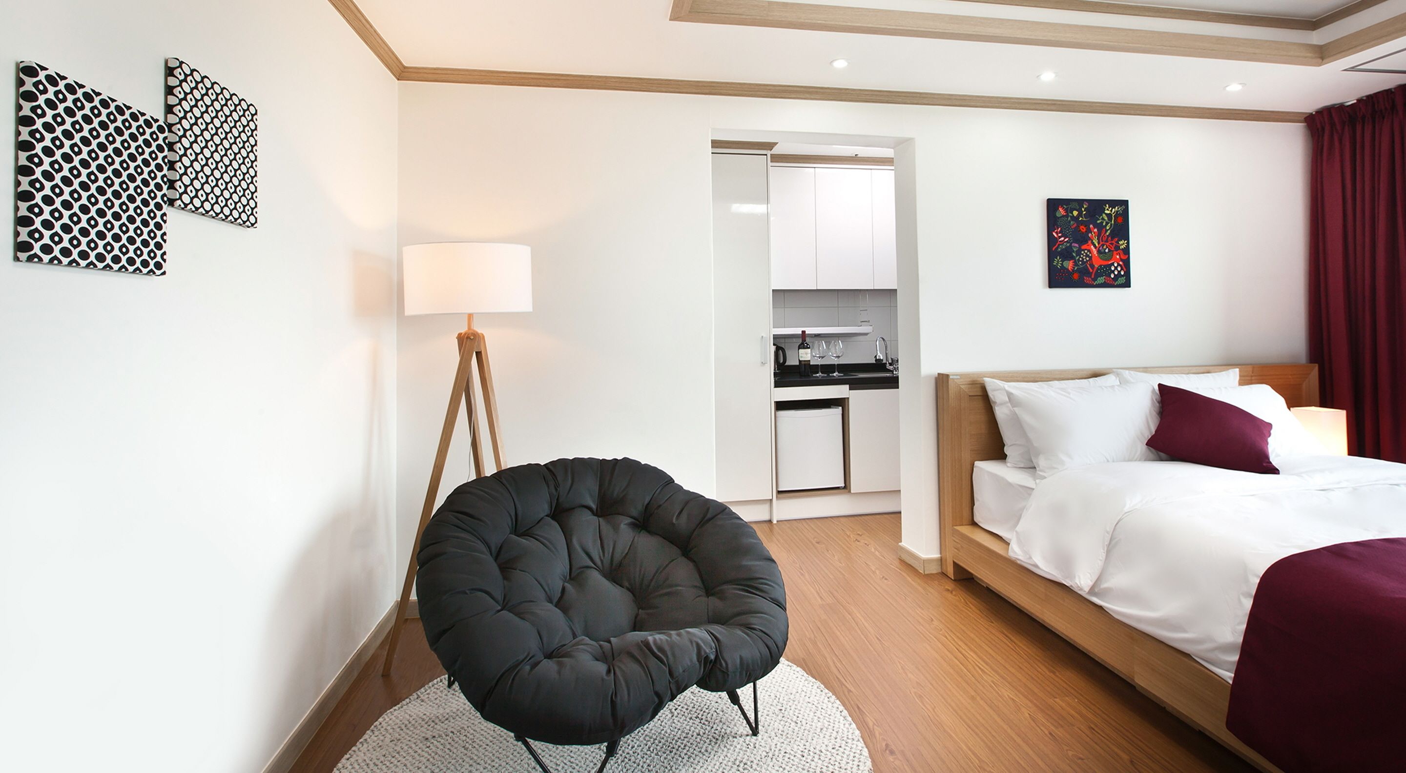 A modern hotel room with a bed, a black armchair, a desk with a lamp, and a kitchen area visible through an open door.