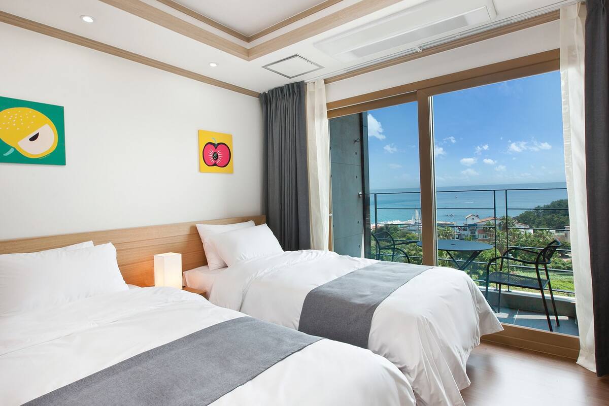 Une chambre d’hôtel avec deux lits, une grande fenêtre donnant sur la mer et des œuvres d’art accrochées aux murs.