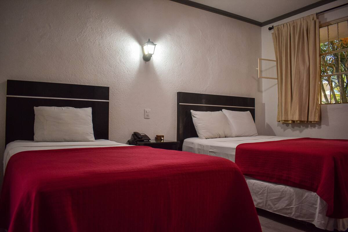 A hotel room with two beds, each with a red bedspread and white pillows, a nightstand with a phone, and a wall-mounted light fixture.