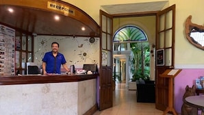 A reception area with a receptionist, a reception desk, and a view of a hallway leading to a room with a large window and a plant. - Hotel Santa Ana (Mérida)