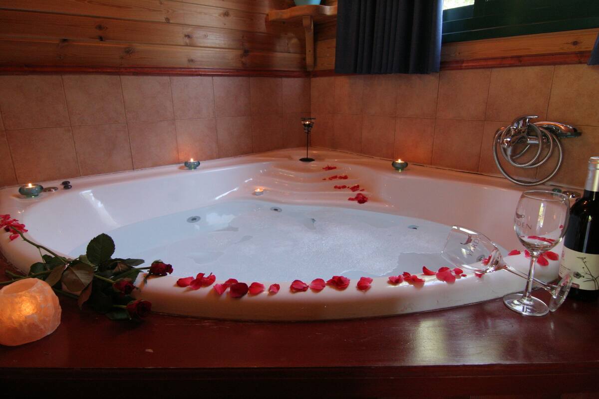 Une baignoire remplie de pétales de rose et entourée de bougies, un verre de vin et une bouteille de vin posés sur une surface en bois.