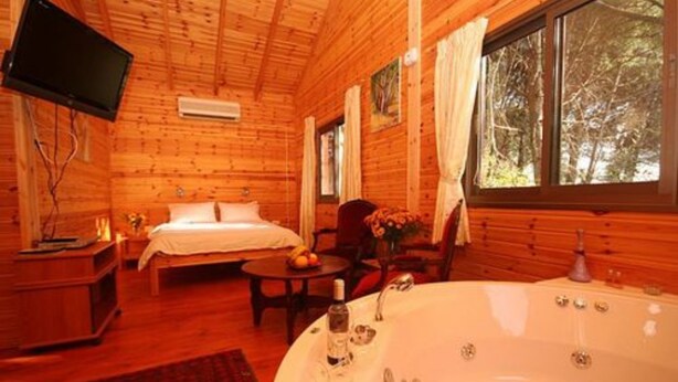 A wooden cabin room with a bed, a sitting area, a large bathtub, and a television mounted on the wall. - Amirim Atmosphere (Merom HaGalil)