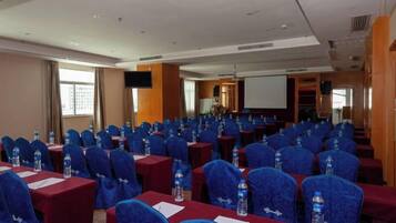 A conference room with rows of tables and chairs, equipped with water bottles and papers, and a projector screen at the front.