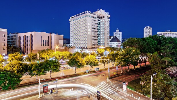 Un hotel moderno de gran altura, con varios pisos y un diseño arquitectónico contemporáneo, rodeado de árboles y una calle urbana bien iluminada.