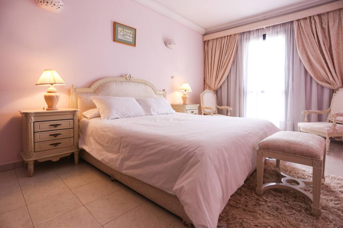 A bedroom with a large bed, two bedside tables with lamps, a chair, and a window with curtains.