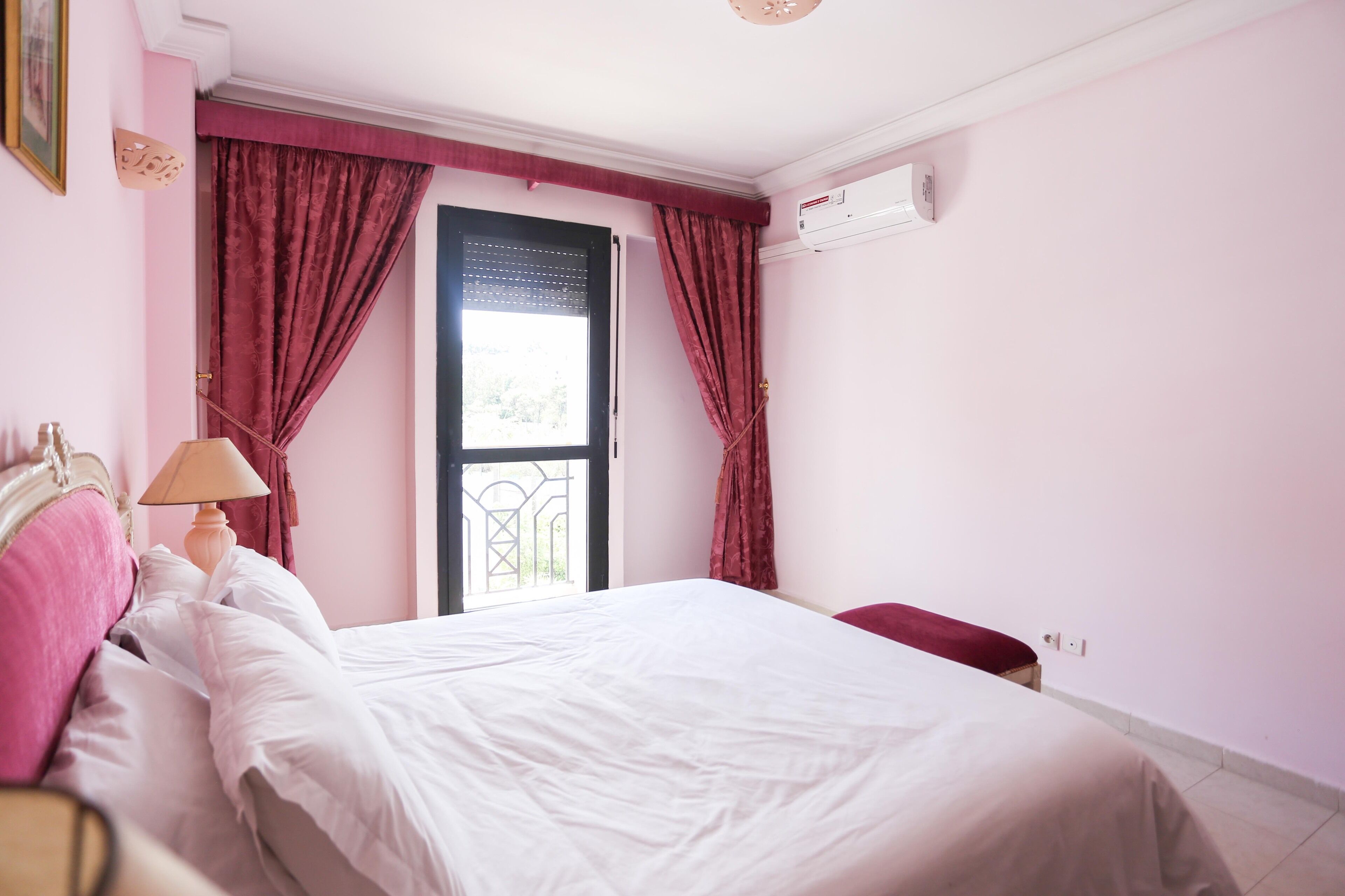 Un dormitorio con cama, mesita de noche, lámpara, ventana con cortinas y equipo de aire acondicionado.