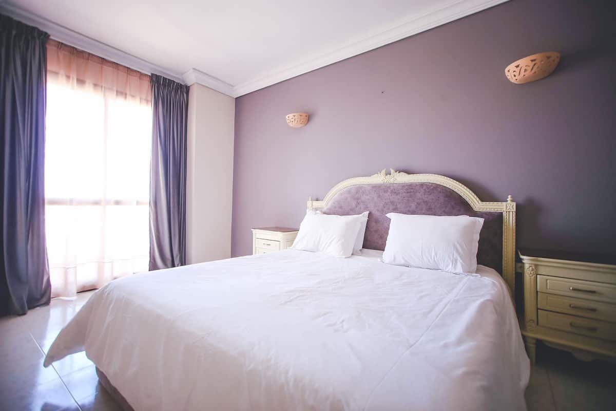 A neatly made bed with white linens, a headboard with a decorative frame, two bedside tables with drawers, and a window with curtains.