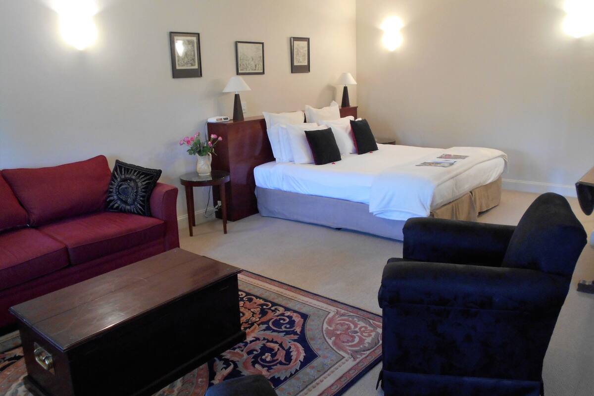 A hotel room with a bed, a red sofa, a dark armchair, a small table with a vase of flowers, and a patterned rug.