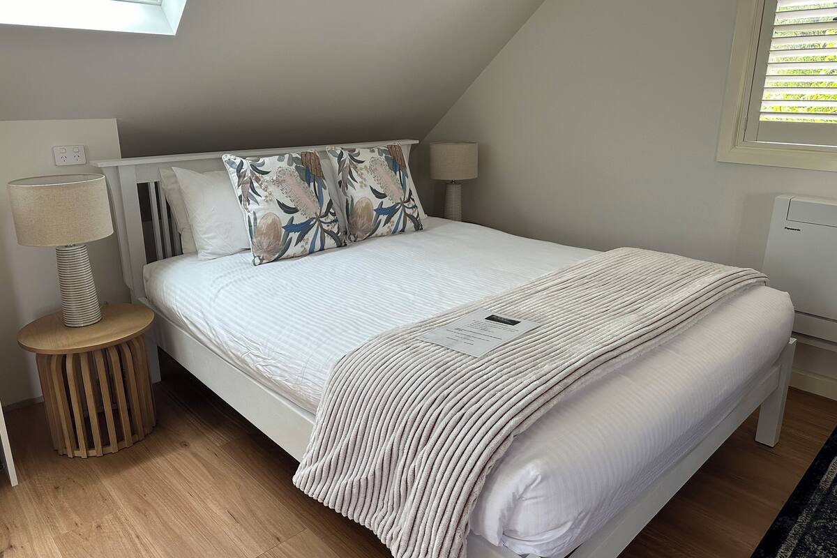 A bedroom with a bed, two bedside lamps, a bedside table, a power outlet, and a window with blinds.