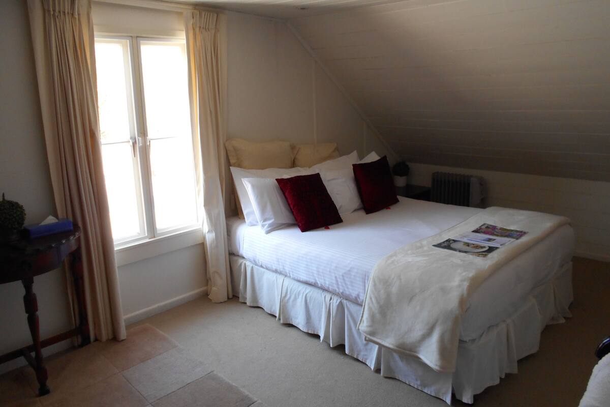 A bedroom with a large bed, a window with curtains, a radiator, and a small table with a plant.