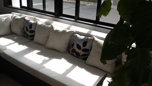 Un área de descanso iluminada por el sol, con sofá, cojines, ventanas amplias y una planta en maceta.