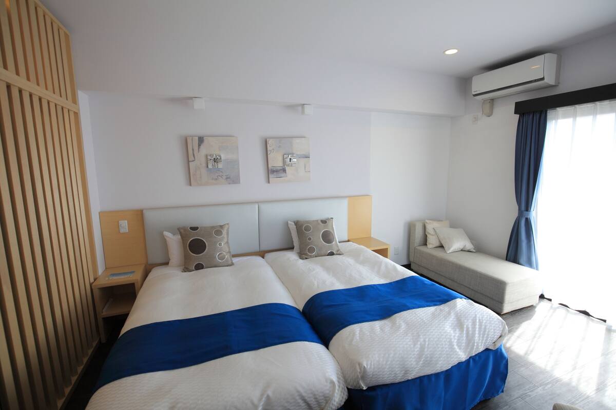 A hotel room with two beds, a sofa, a wall-mounted air conditioner, and artwork on the wall.