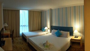 Ein Hotelzimmer mit zwei Betten, einem Schreibtisch und einem Stuhl. Vor den Fenstern hängen Vorhänge und auf dem Schreibtisch steht eine Lampe.