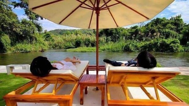 Two wooden lounge chairs with a table and an umbrella, overlooking a river with greenery on both sides. - Worawee Resort & Spa (Kaeng Krachan)