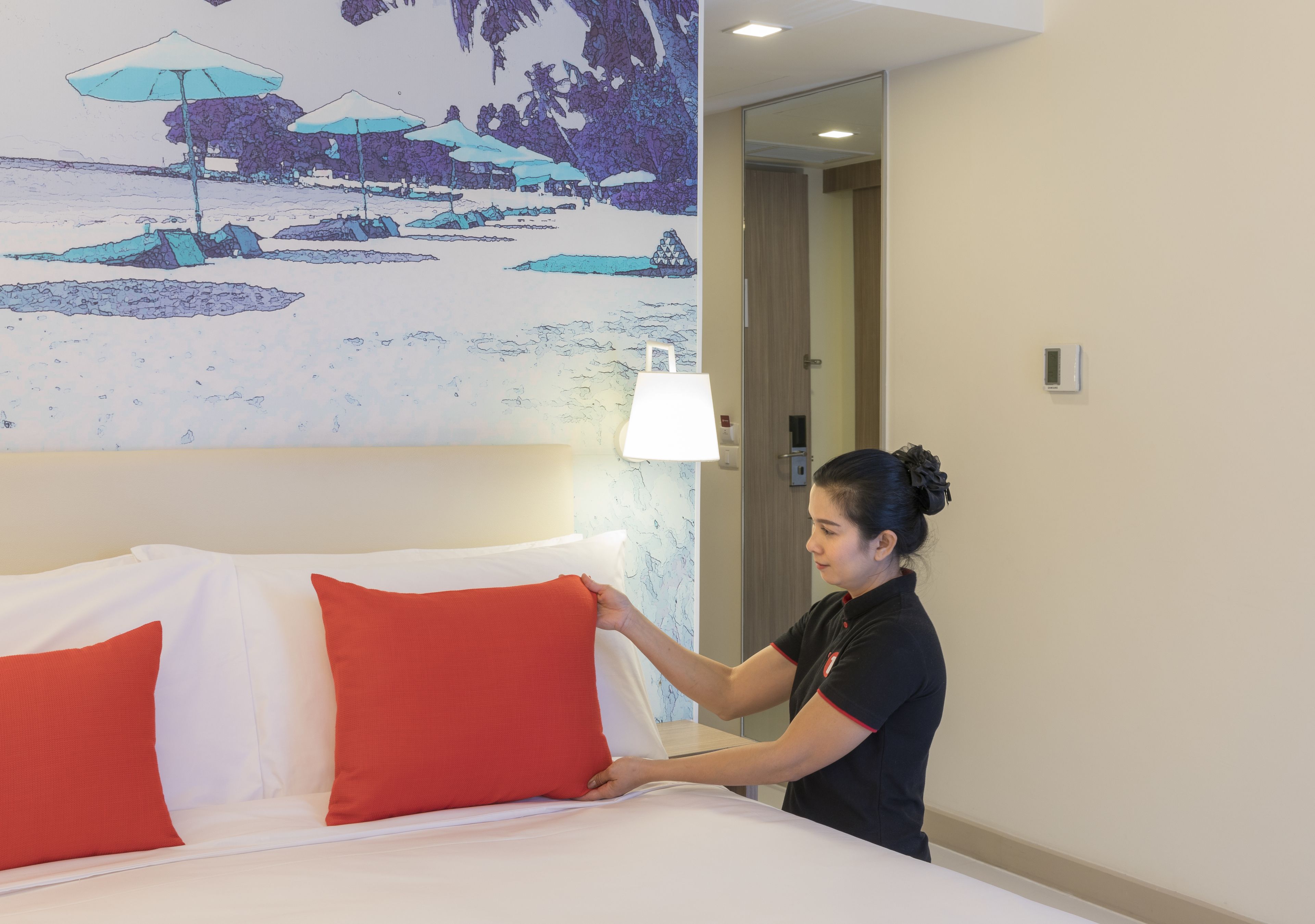 Una habitación de hotel con cama, almohadas, un mural en la pared, una lámpara y una puerta.