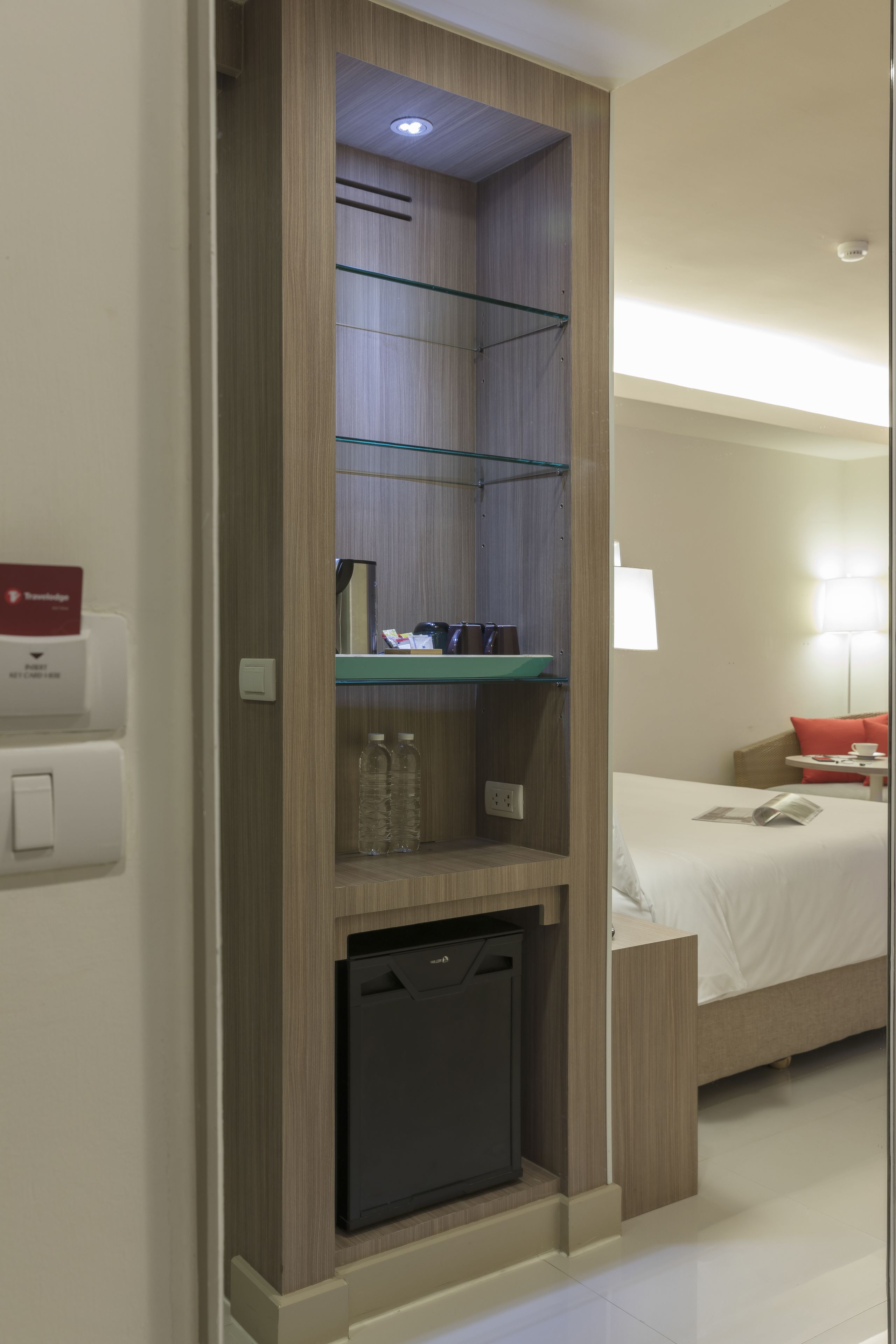 A hotel room with a bed, a small desk, a shelf with toiletries, and a compact refrigerator.