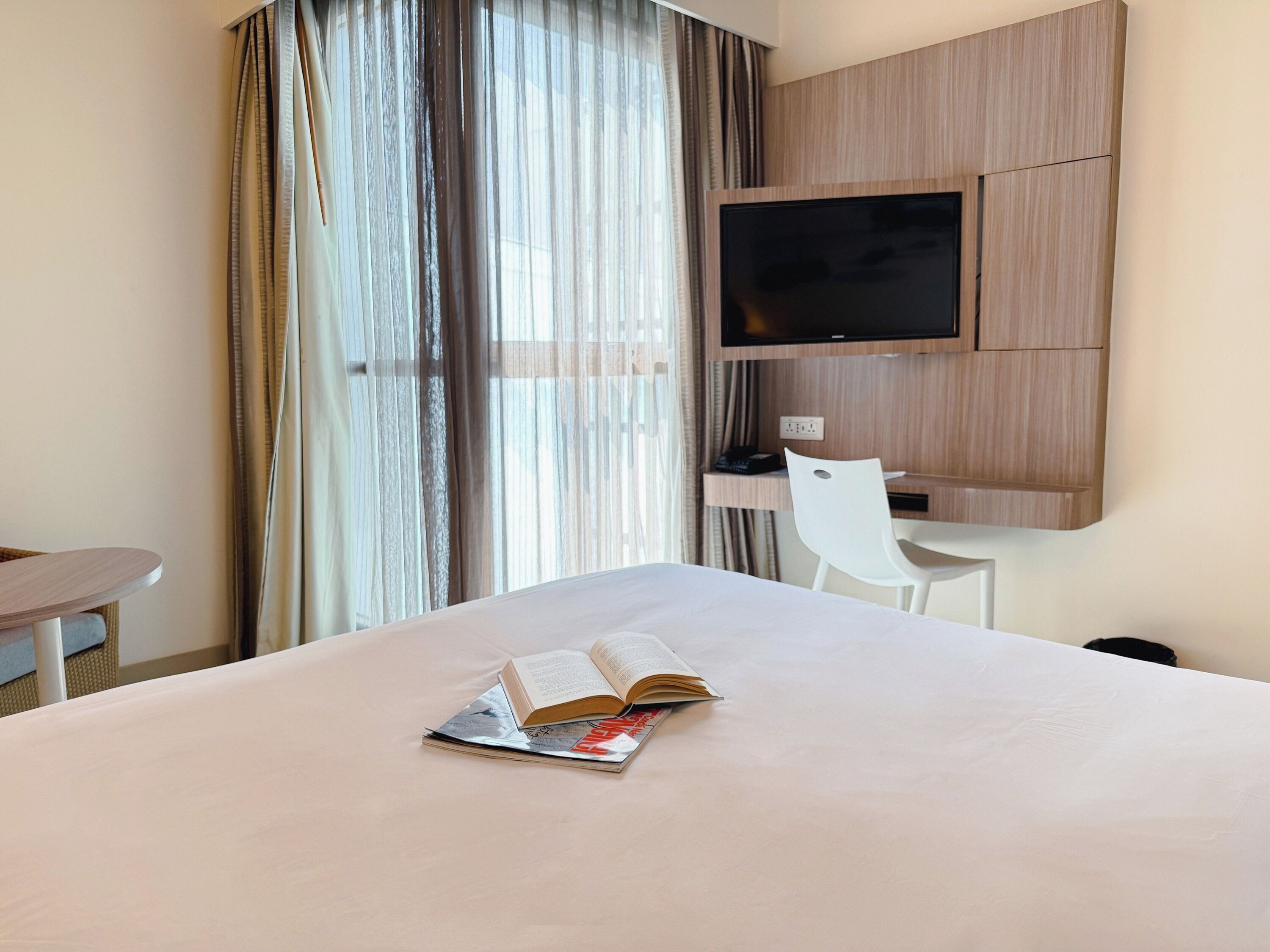 A hotel room with a bed, a desk, a chair, a TV mounted on the wall, and a window with curtains.