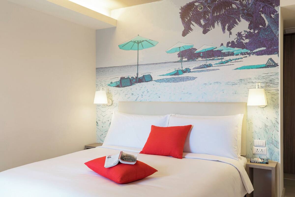 Une chambre d’hôtel avec un lit, deux oreillers rouges, un livre et une fresque murale représentant une scène de plage.