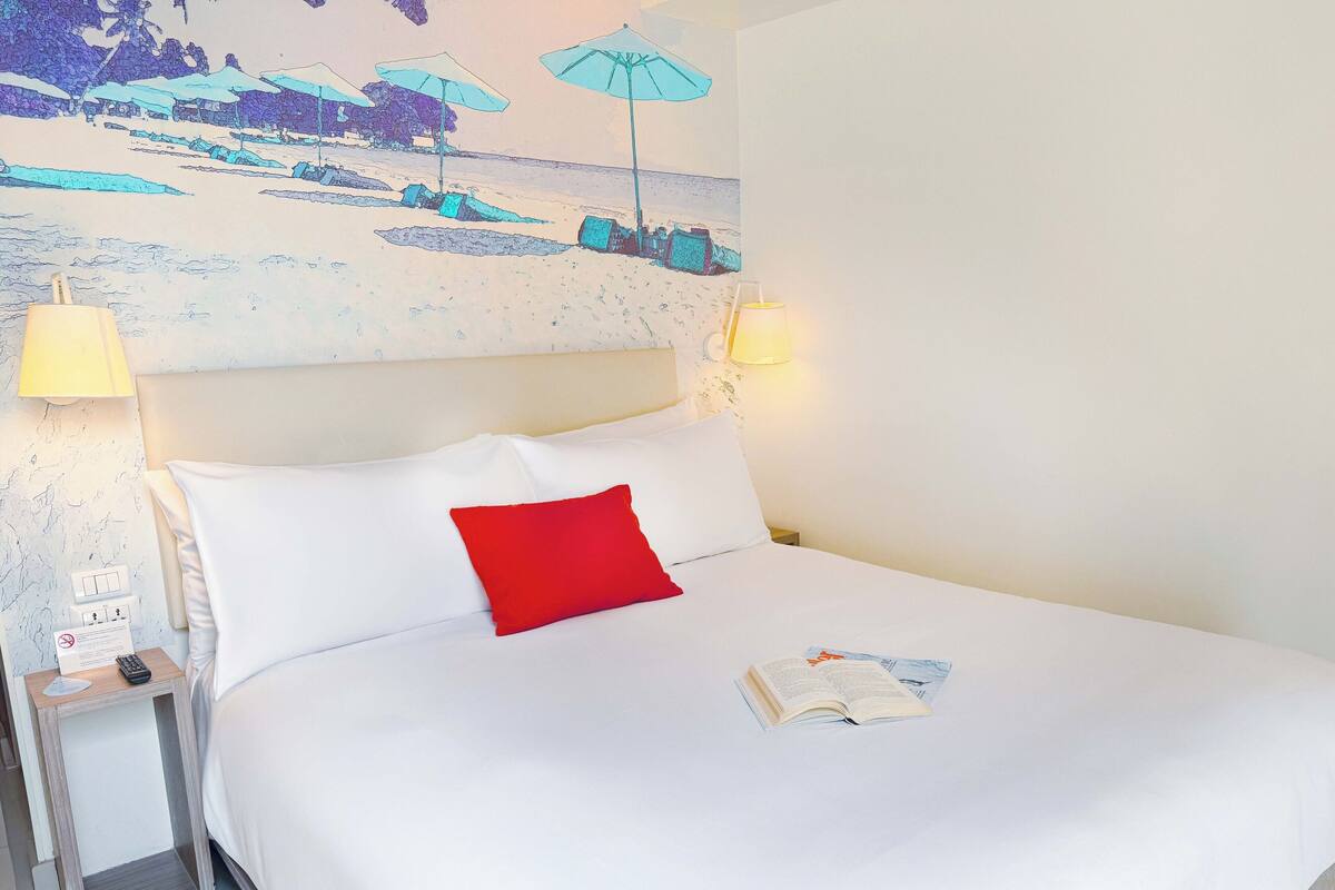 Une chambre d’hôtel avec un lit, une table de chevet avec une lampe, un tableau au mur et une fenêtre.