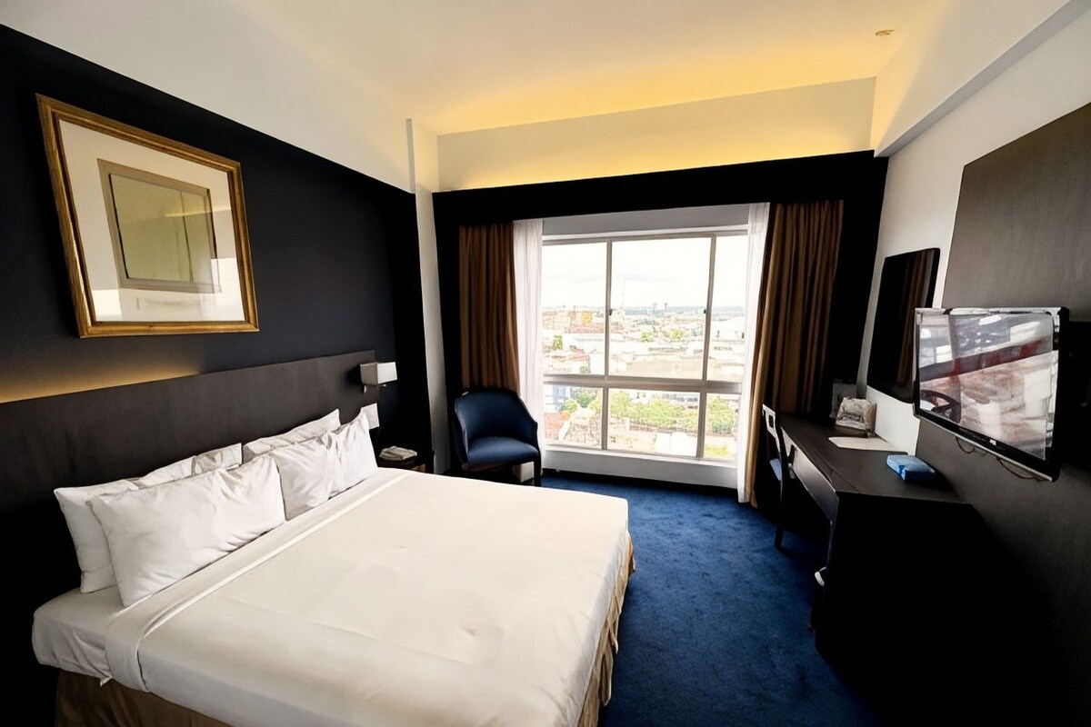 A hotel room with a large bed, a desk with a computer, a chair, a TV, and a window with curtains.