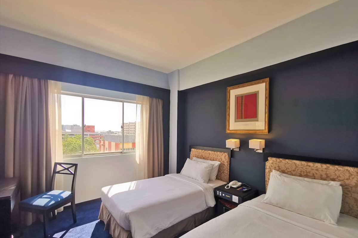Ein Hotelzimmer mit zwei Betten, einem Schreibtisch, einem Stuhl, einem Fenster mit Vorhängen, einem gerahmten Bild an der Wand und einem Nachttisch mit Telefon und Lampe.