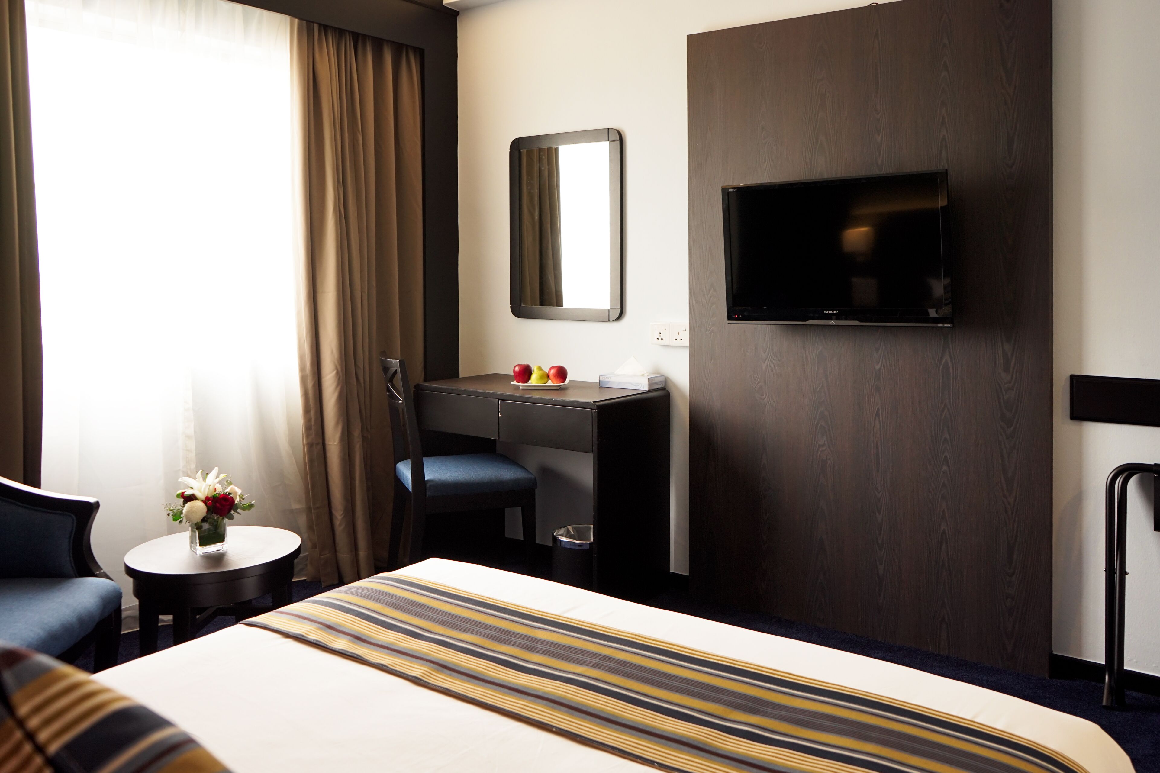 Habitació d'hotel amb un llit, escriptori, cadira, taula petita amb un vas de flors, televisió penjada a la paret i finestra amb cortines.