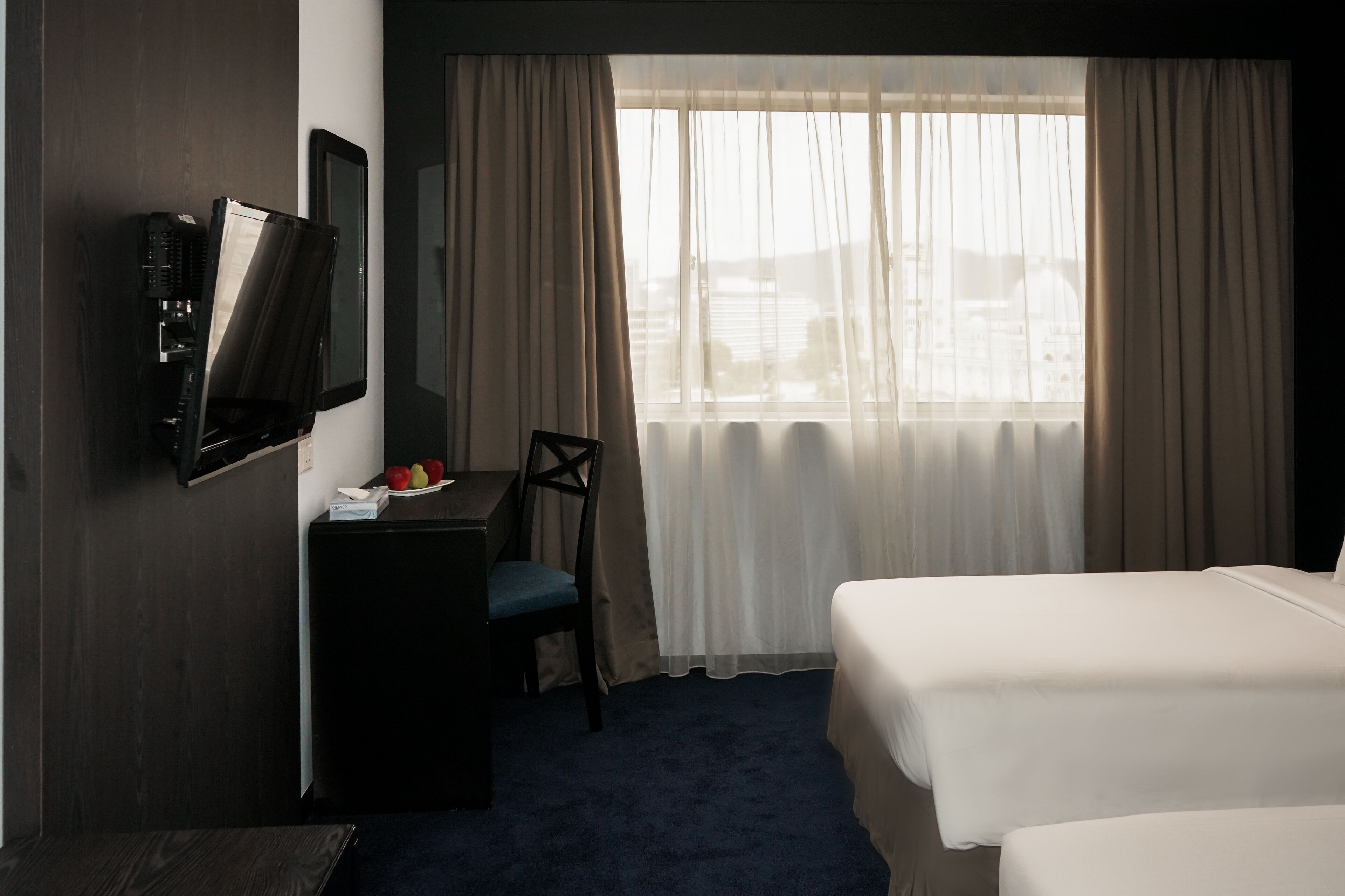 A hotel room with a bed, a desk with a chair, a television mounted on the wall, and a window with curtains.