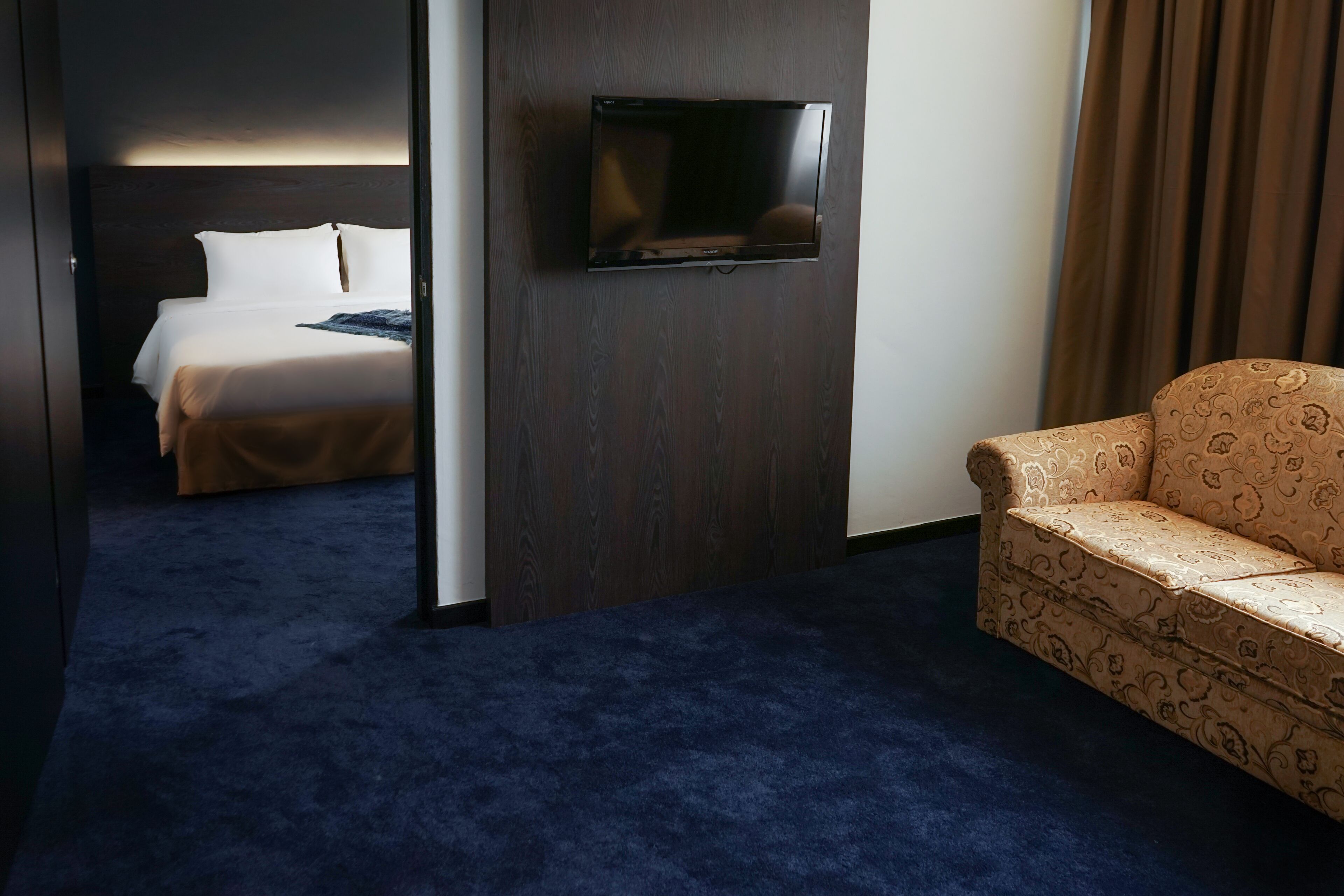 A hotel room with a bed, a sofa, a TV mounted on the wall, and a dark blue carpet.
