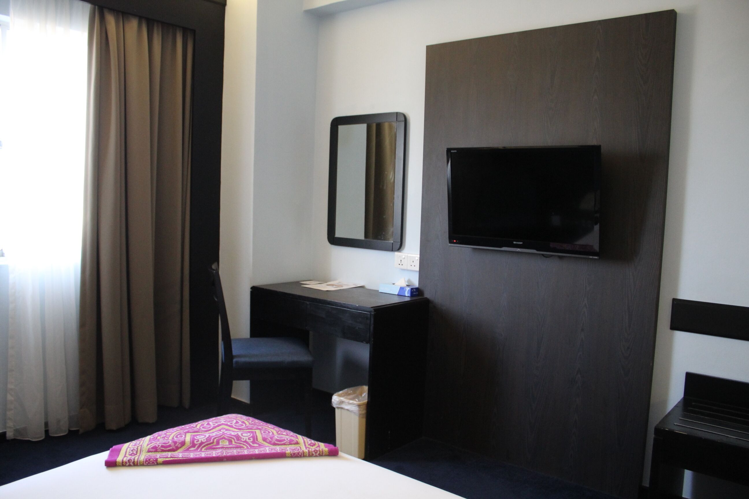 A hotel room with a bed, a desk, a chair, a television mounted on the wall, and a window with curtains.