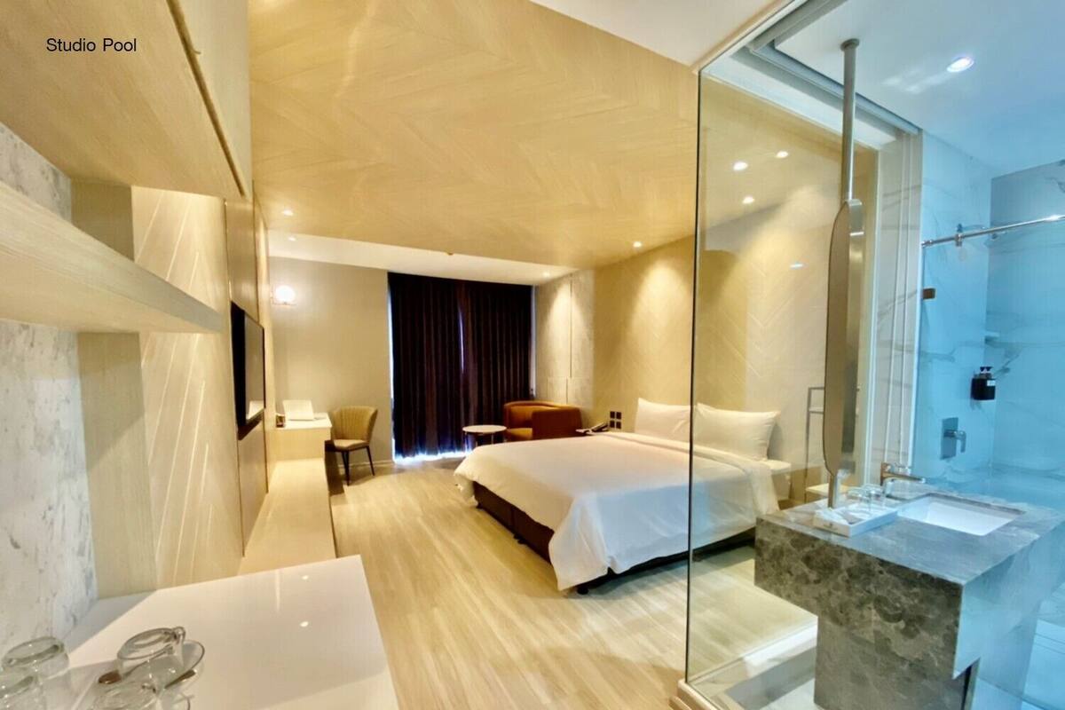 A modern hotel room with a large bed, a glass-enclosed shower, a sitting area with a chair and table, and a small desk with a laptop.