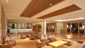 Um hall amplo com design moderno, destacando uma área de estar central com sofás e poltronas, um balcão de recepção e uma grande parede de vidro com vista para o interior.