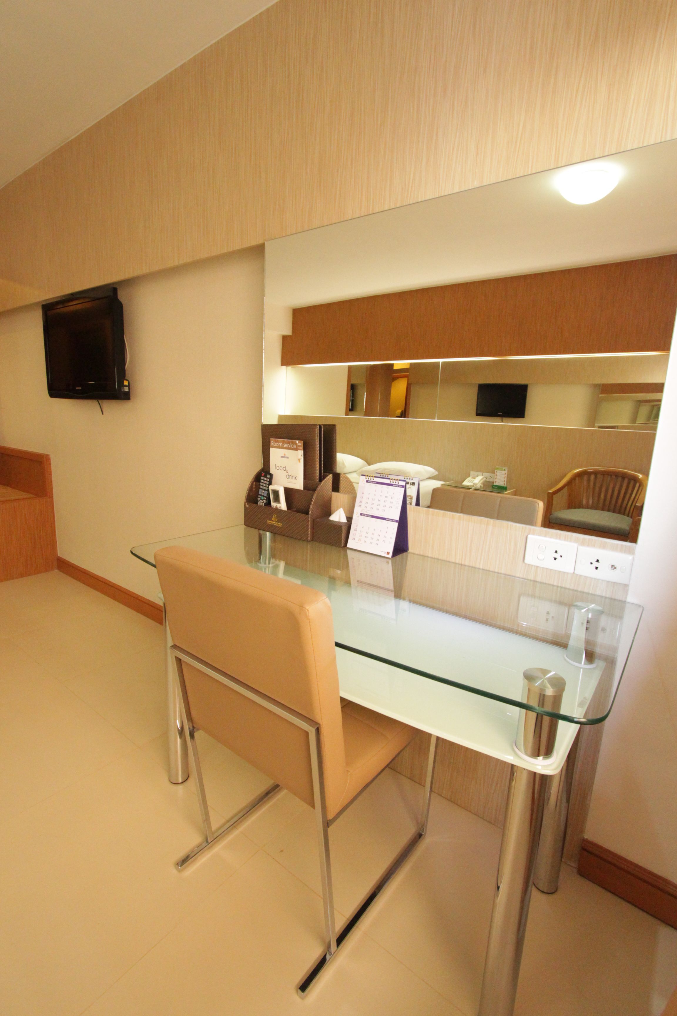 A modern hotel room with a glass dining table, a cushioned chair, a flat-screen TV mounted on the wall, a bedside table with a lamp, and a built-in shelf with decorative items.