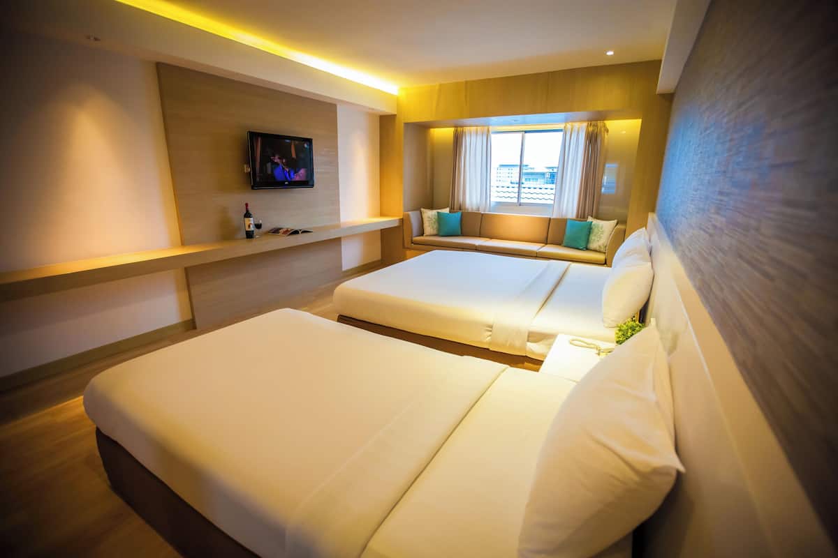 A hotel room with a large bed, a TV mounted on the wall, a window with curtains, and a sofa with cushions.