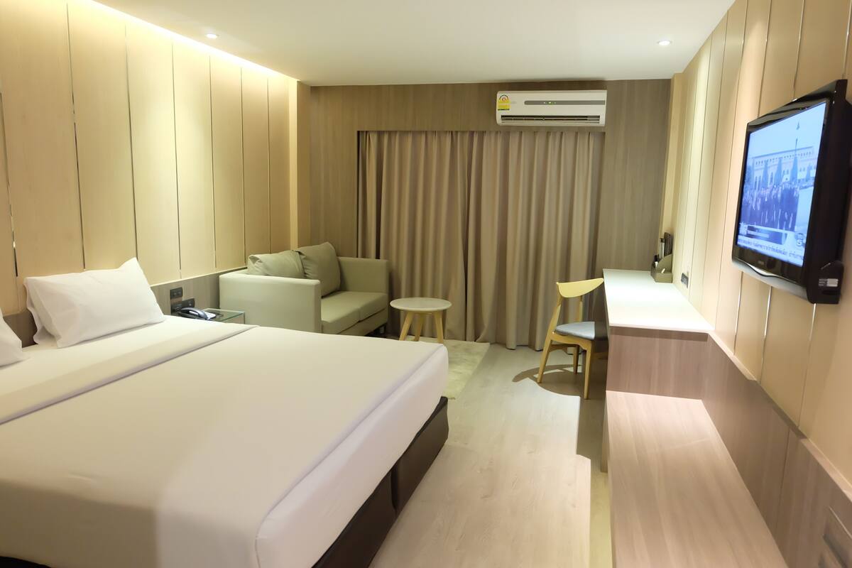 A hotel room with a large bed, a sofa, a small table, a TV, and an air conditioning unit.