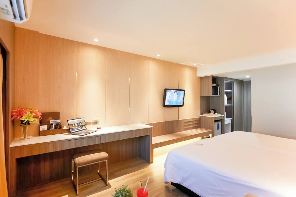 A modern hotel room with a wooden panel wall, a flat-screen TV mounted above a desk, a bed with white bedding, and a small seating area.
