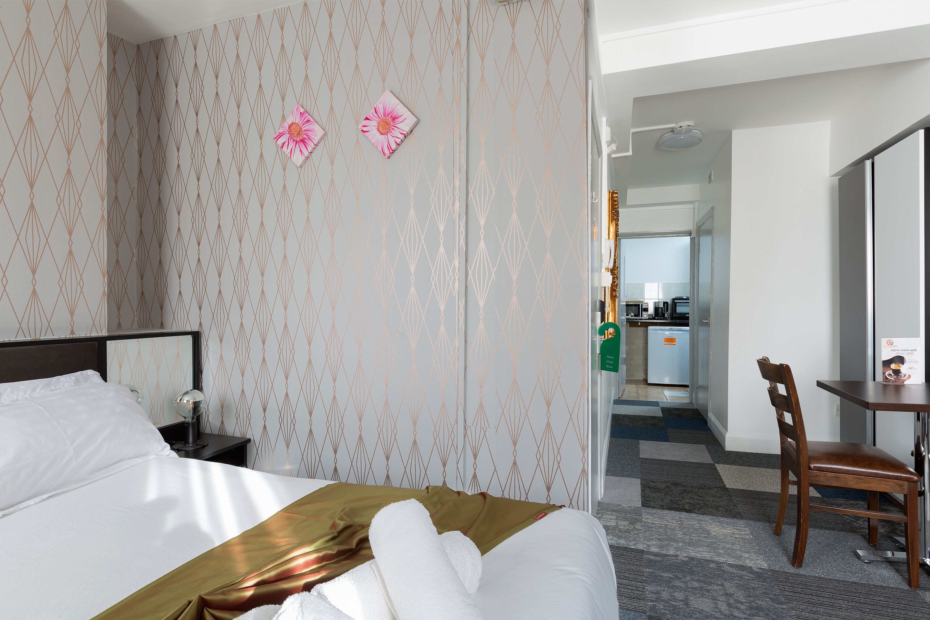 A hotel room with a bed, a nightstand, a desk, and a chair. There is a kitchen area visible in the background.