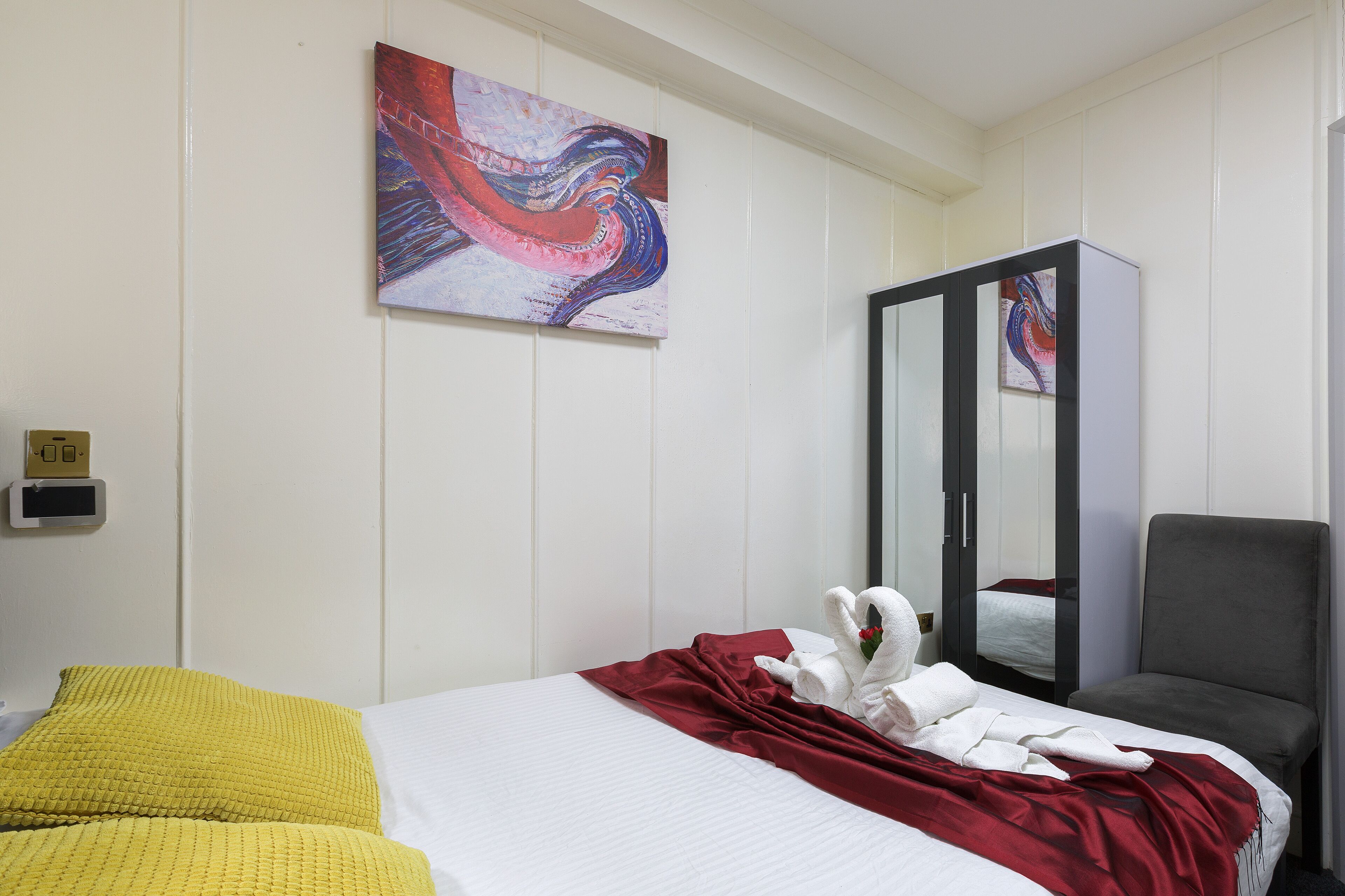 Habitación de hotel con cama, una colcha roja, almohada amarilla y un sillón gris, además de un cuadro en la pared.