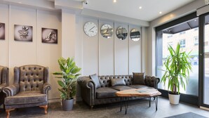A modern lobby with a leather sofa, a tufted armchair, and a coffee table. There are four wall clocks and a large potted plant. - Central Hoxton Shoreditch (London)