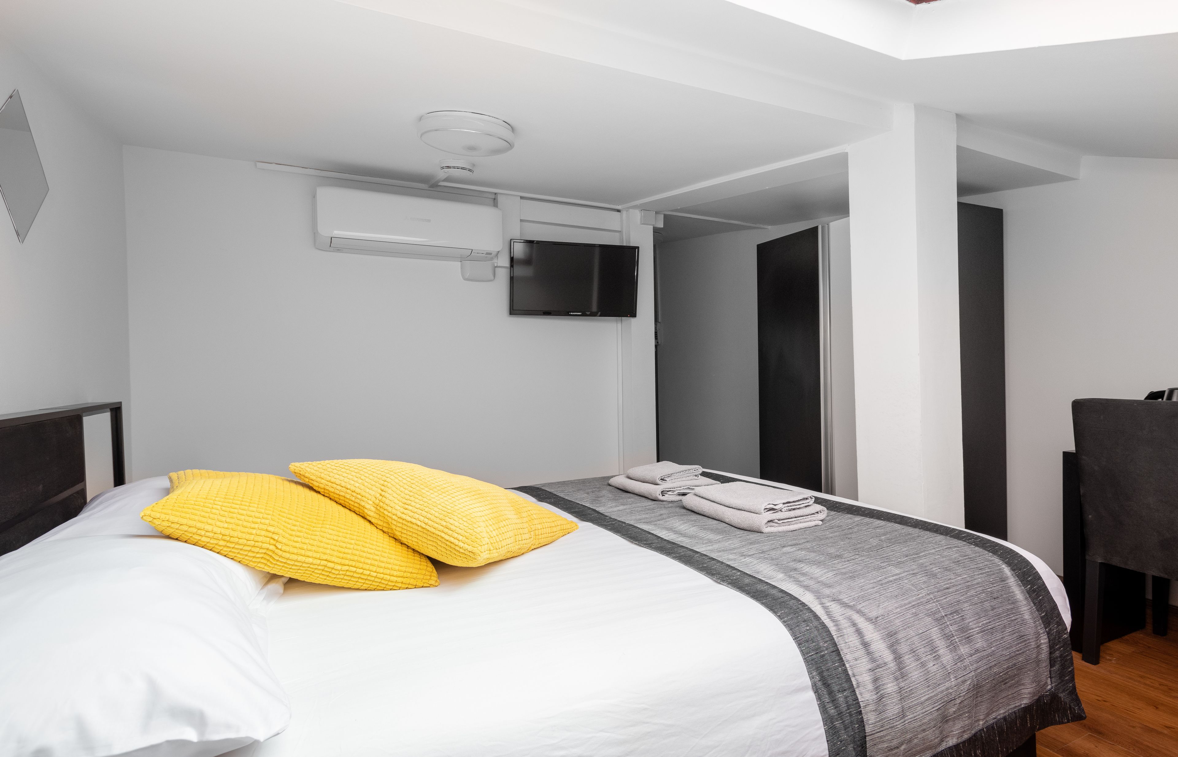 A modern hotel room with a neatly made bed, a flat-screen TV mounted on the wall, and a visible air conditioning unit.