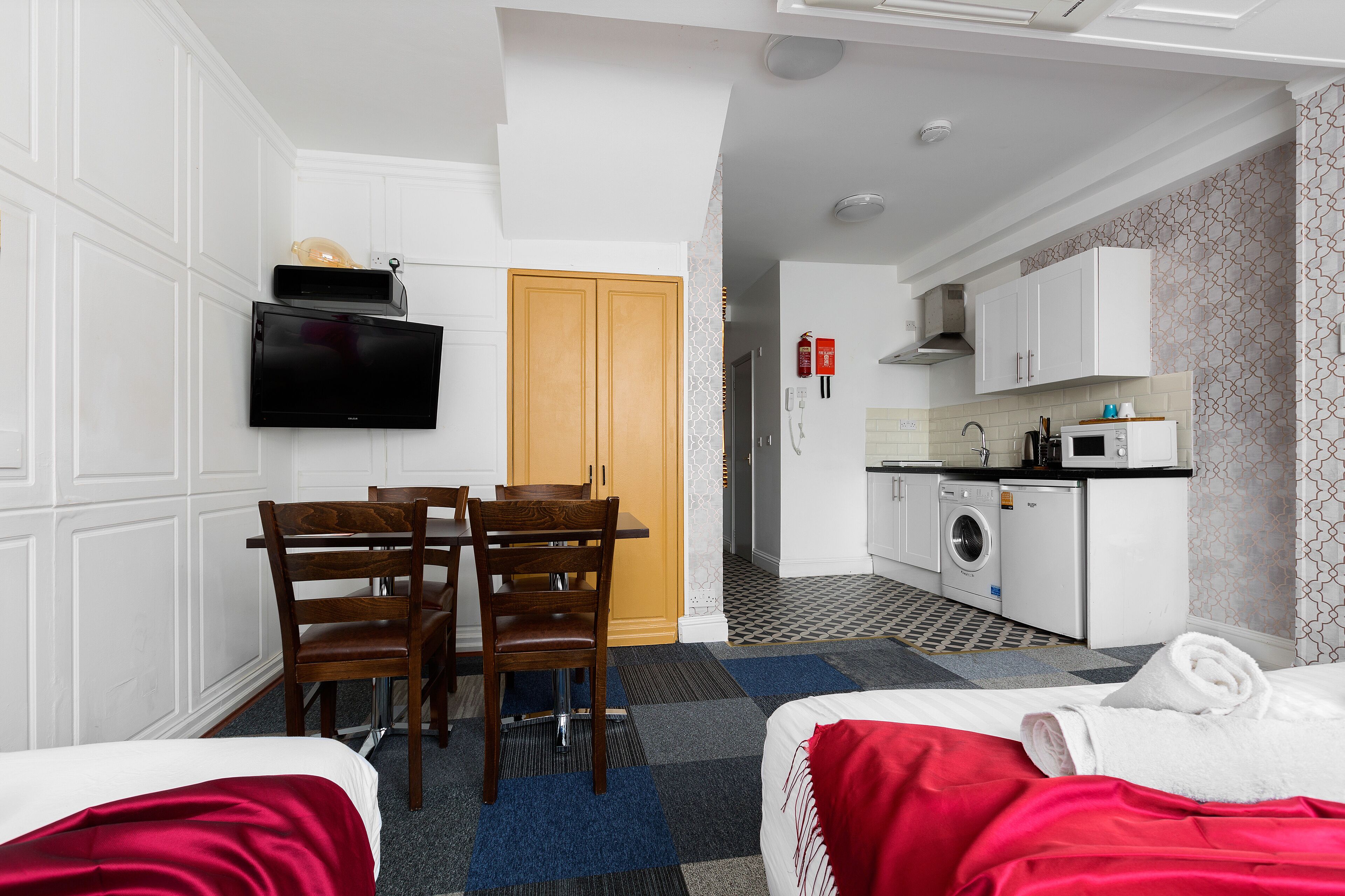 A hotel room with a bed, a desk with chairs, a television, a washing machine, and a kitchenette.