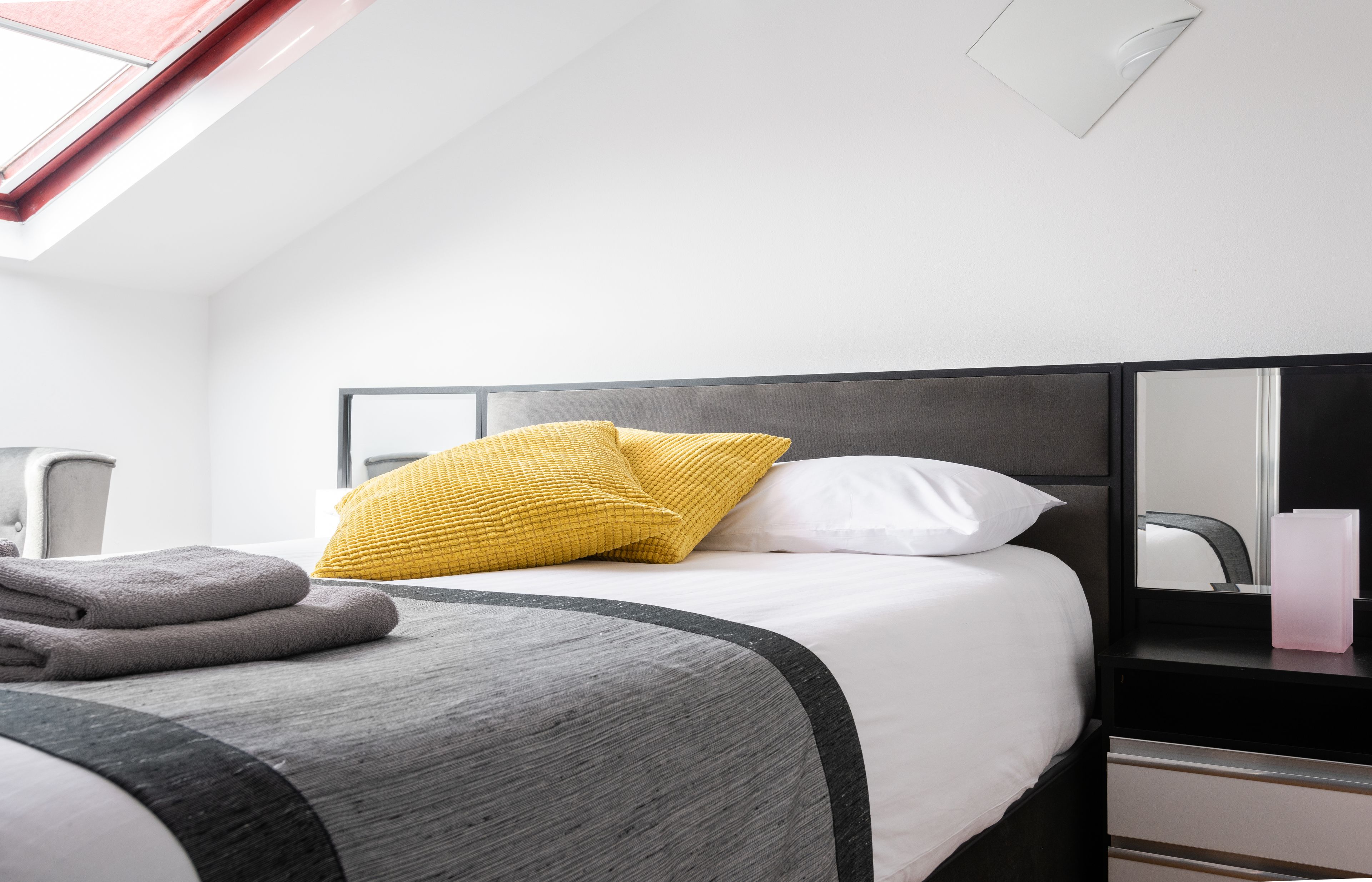 A hotel room with a bed, a bedside table, a mirror, and a skylight.