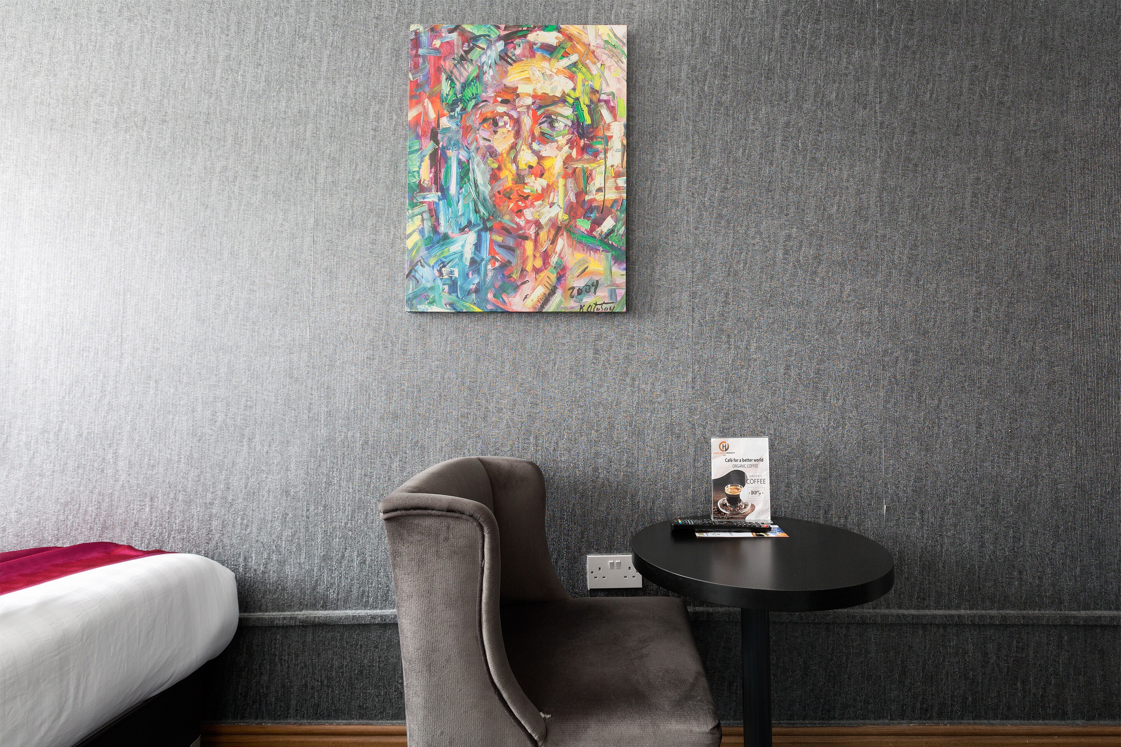 A hotel room with a grey textured wall, a grey armchair, a small round table with a remote control and a brochure, and a bed with a red blanket.