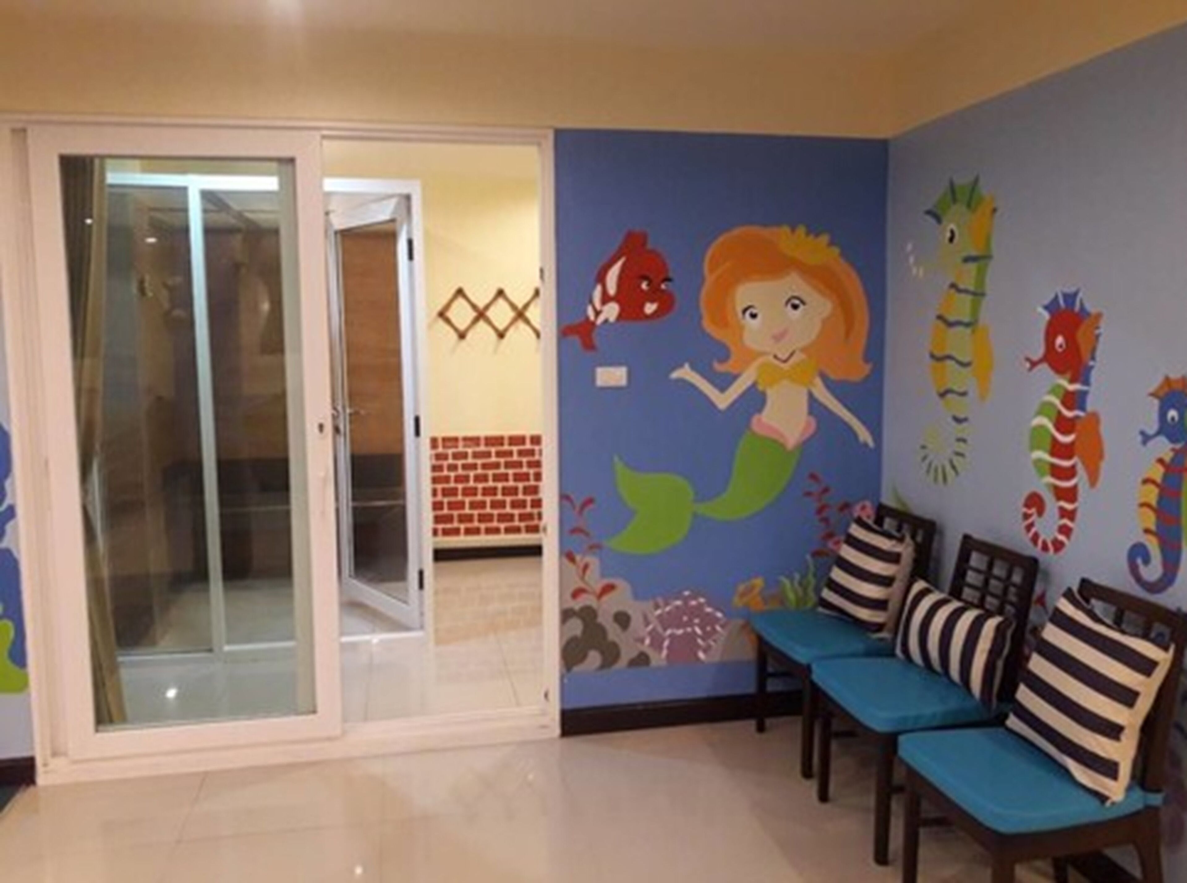 A room with a blue wall mural featuring a mermaid and seahorses, a glass door leading to a tiled area, and two blue cushioned chairs with striped pillows.