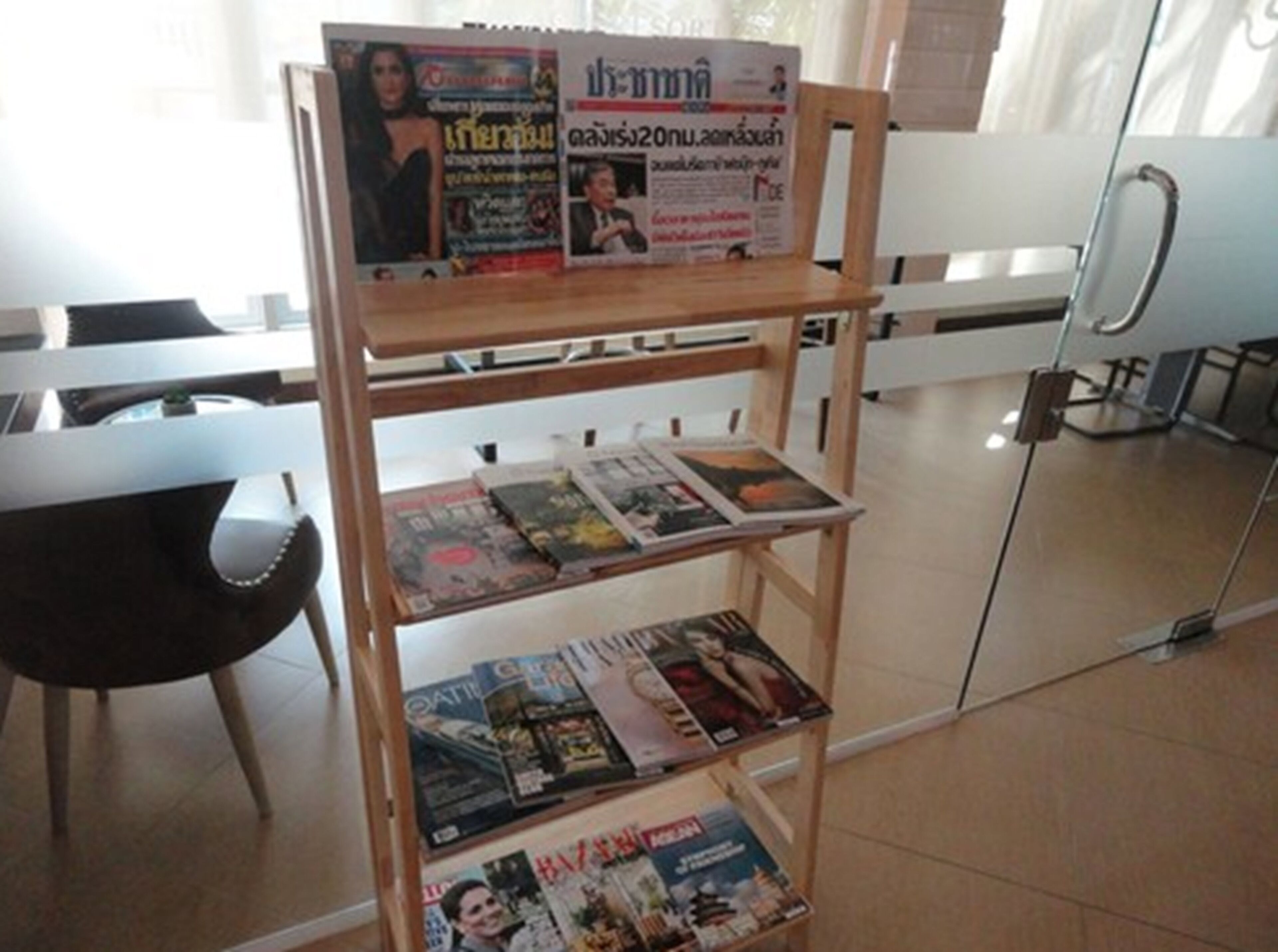 Um suporte de madeira para revistas com várias revistas, uma cadeira e uma porta de vidro ao fundo.